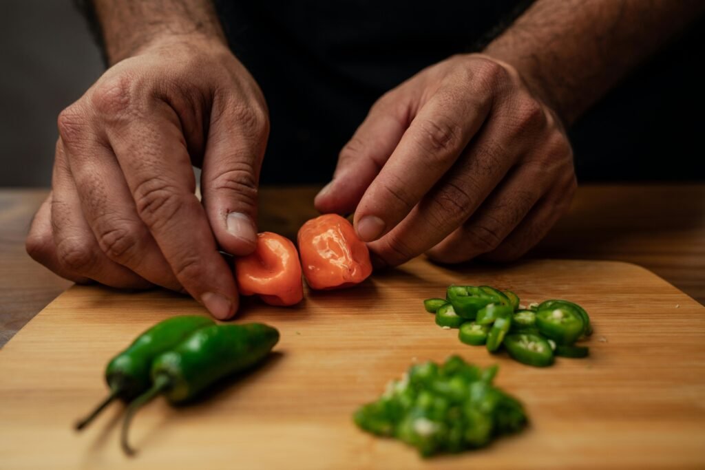 Close-up of hands slicing jalapeno and ghost peppers on a wooden chopping board.